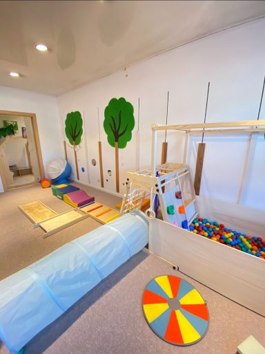 Spielzimmer mit bunten Matten, einem Tunnel und Holzspielzeugen. Grüne Bäume an der Wand.