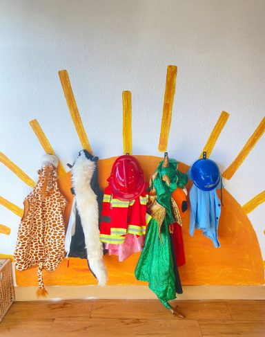Fünf Kostüme in verschiedenen Farben hängen an einer Wand, die eine Sonnenscheibe darstellt.