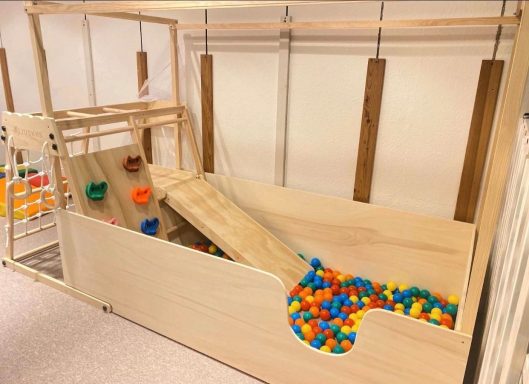 Holzspielplatz mit Rutsche und bunten Bällen in einem sicheren Bereich.